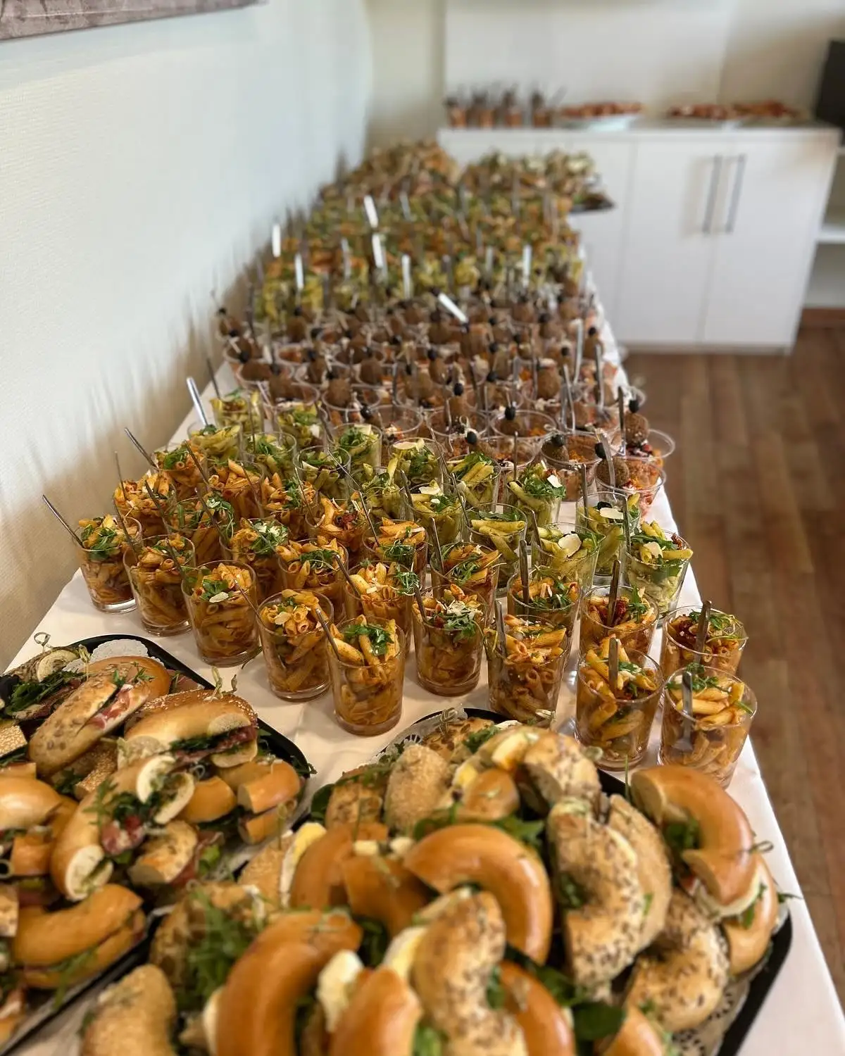 Long catering table with pasta cups in glasses and platters of filled bagels