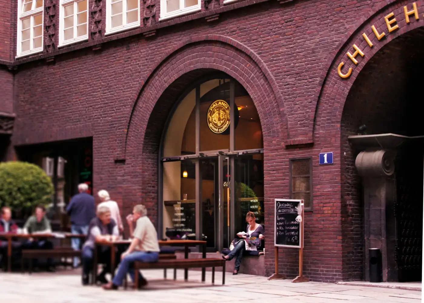 Entrance of PALANG good coffee in the Chilehaus: guests at tables in front of the brick arch, the logo in the window